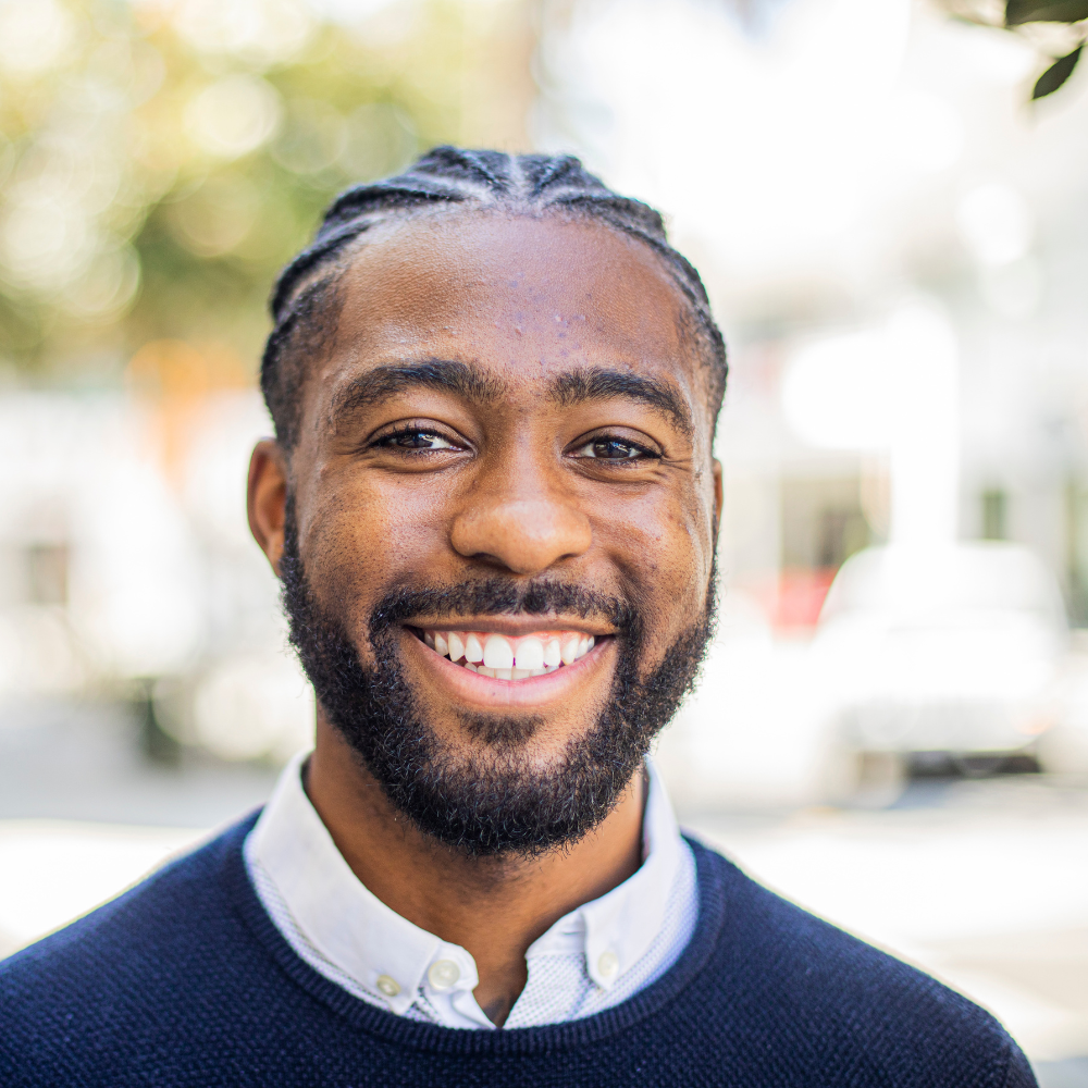Portrait of a smiling man with braided hair and a beard, wearing a dark sweater over a white collared shirt outdoors.