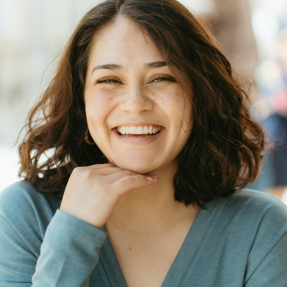 Smiling woman with medium curly hair resting her chin on her hand wearing a teal top.