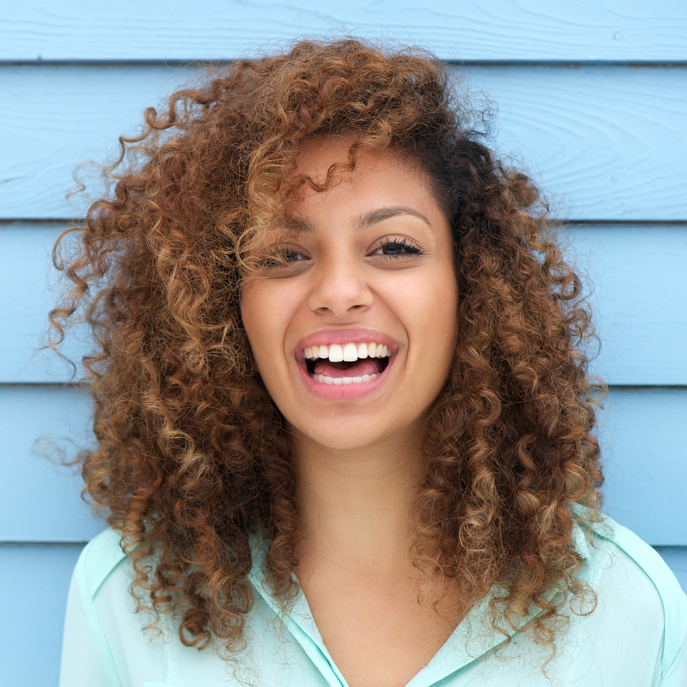 Smiling young woman with curly hair wearing a light blue shirt against a blue wooden background.