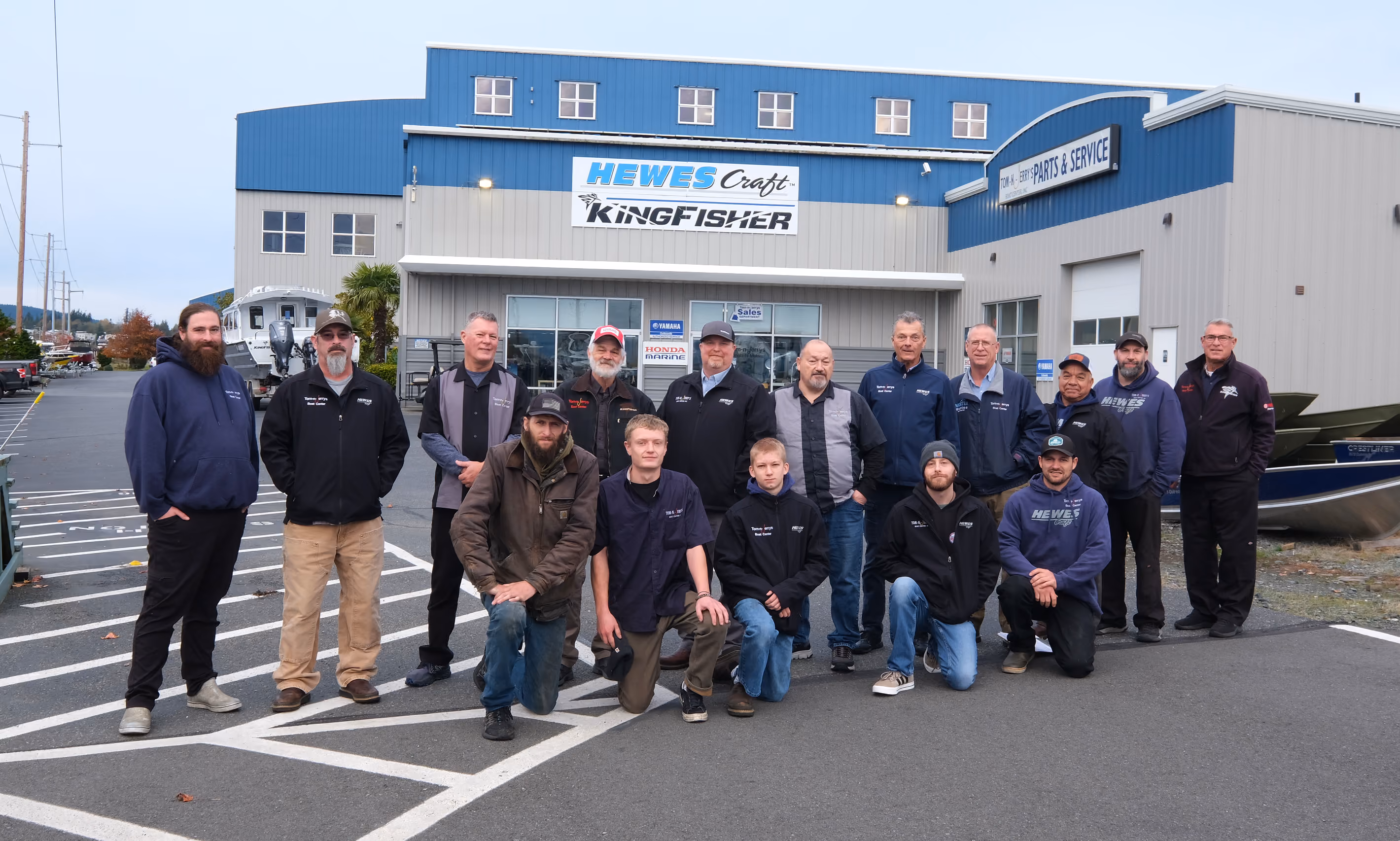 Group of 15 men posing outside a blue and gray building with signs for Hewes Craft, KingFisher, and Tom-n-Jerry Parts & Service.