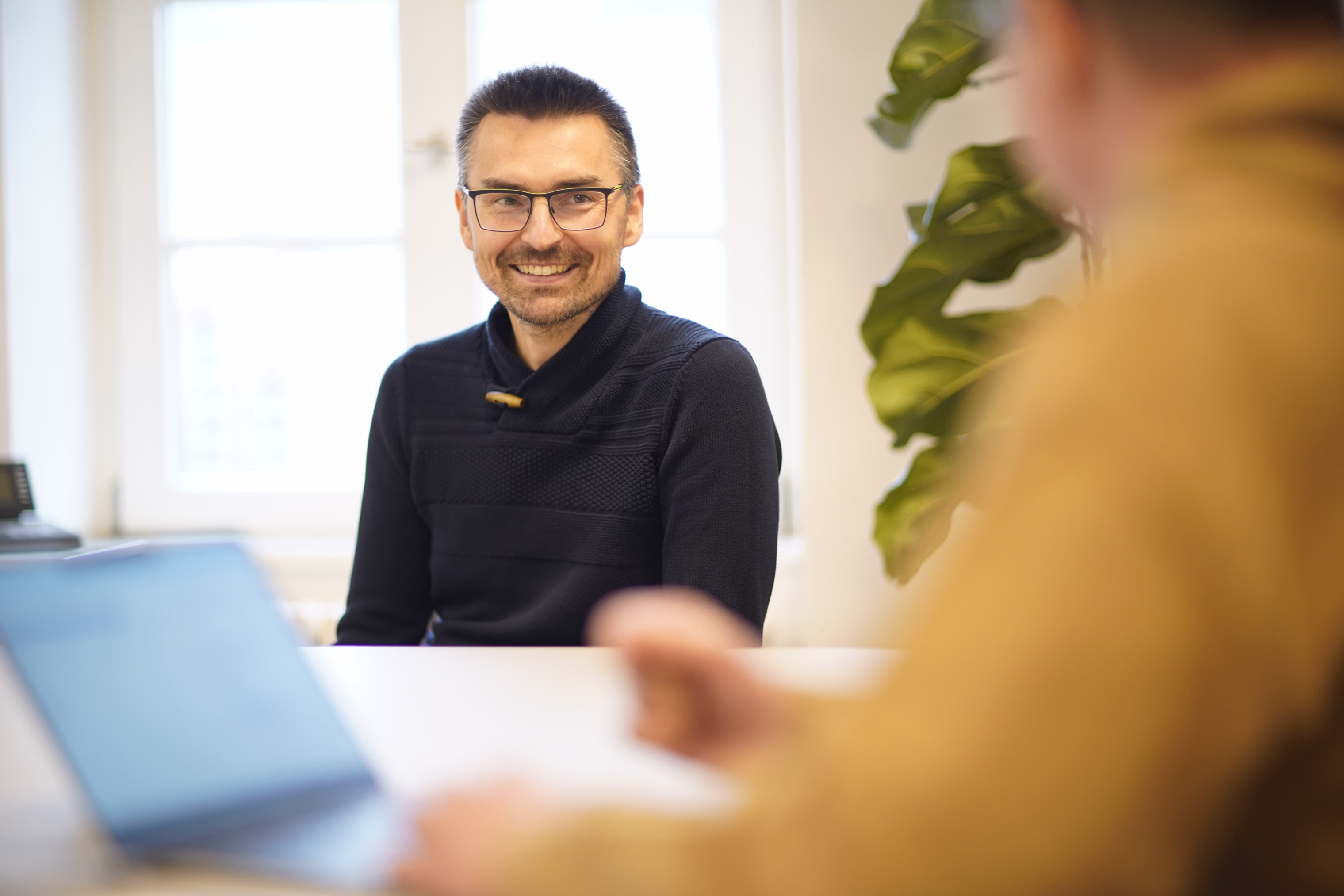 Lächelnder Mann mit Brille in einem schwarzen Pullover sitzt an einem Tisch und unterhält sich mit einer anderen Person mit einem Laptop.
