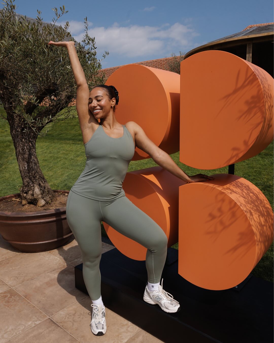 Woman stretching outdoors beside modern orange fitness props, wearing green Sweaty Betty activewear.