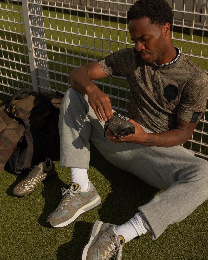 Athlete sitting on turf examining New Balance x Stone Island boots while wearing matching training kit.