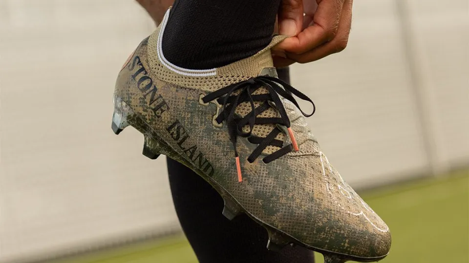 New Balance x Stone Island football boot featuring digital camouflage design and branding detail.