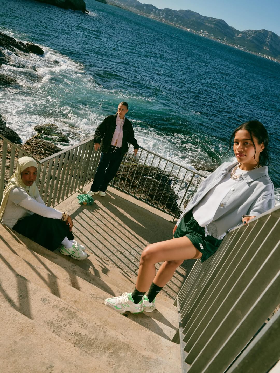 Models wearing New Balance x Foot Locker Club Ivy collection relaxing by the ocean in sporty streetwear and sneakers.
