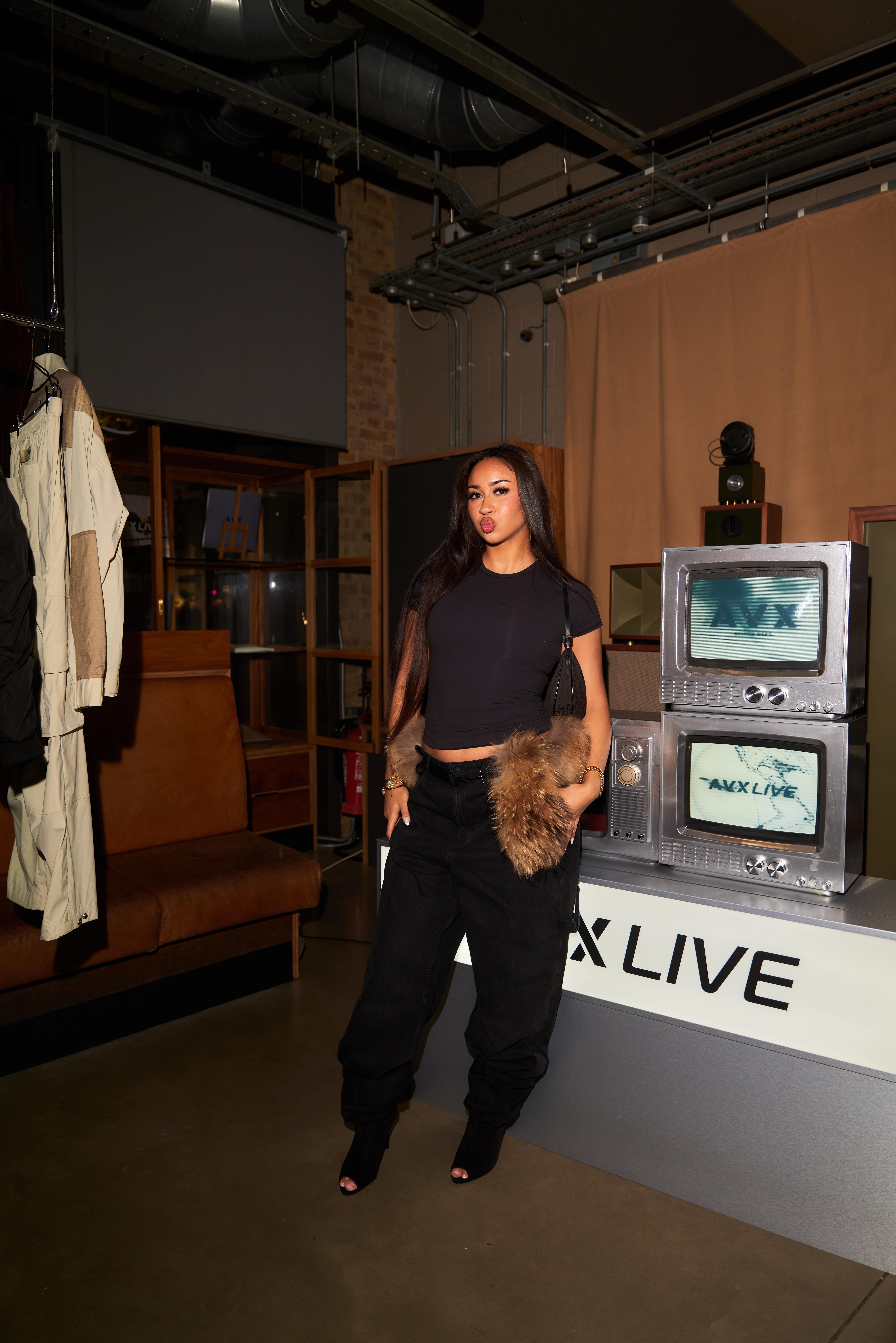 Woman guest wearing black crop top and AVX apparel posing beside vintage TV installation at AVX Live venue.