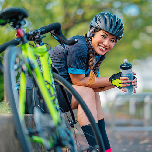 Een jonge, lachende vrouw zit neer en houdt een drinkfles vast terwijl ze een pauze neemt van het fietsen.