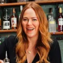 Smiling woman with long wavy red hair sitting in front of a bar with bottles on shelves.