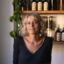 Smiling woman with blonde hair and glasses standing in front of a kitchen shelf with plants and bottles.
