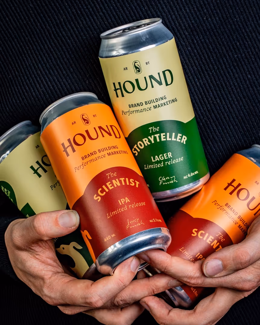 Hands holding four colorful cans of HOUND brand craft beer labeled The Scientist IPA and The Storyteller Hoped Lager.