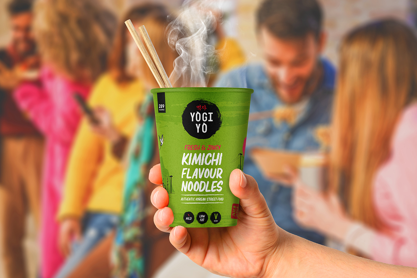 Hand holding a green cup of steaming Yogi Yo kimchi flavour noodles with chopsticks, blurred group of people in the background.