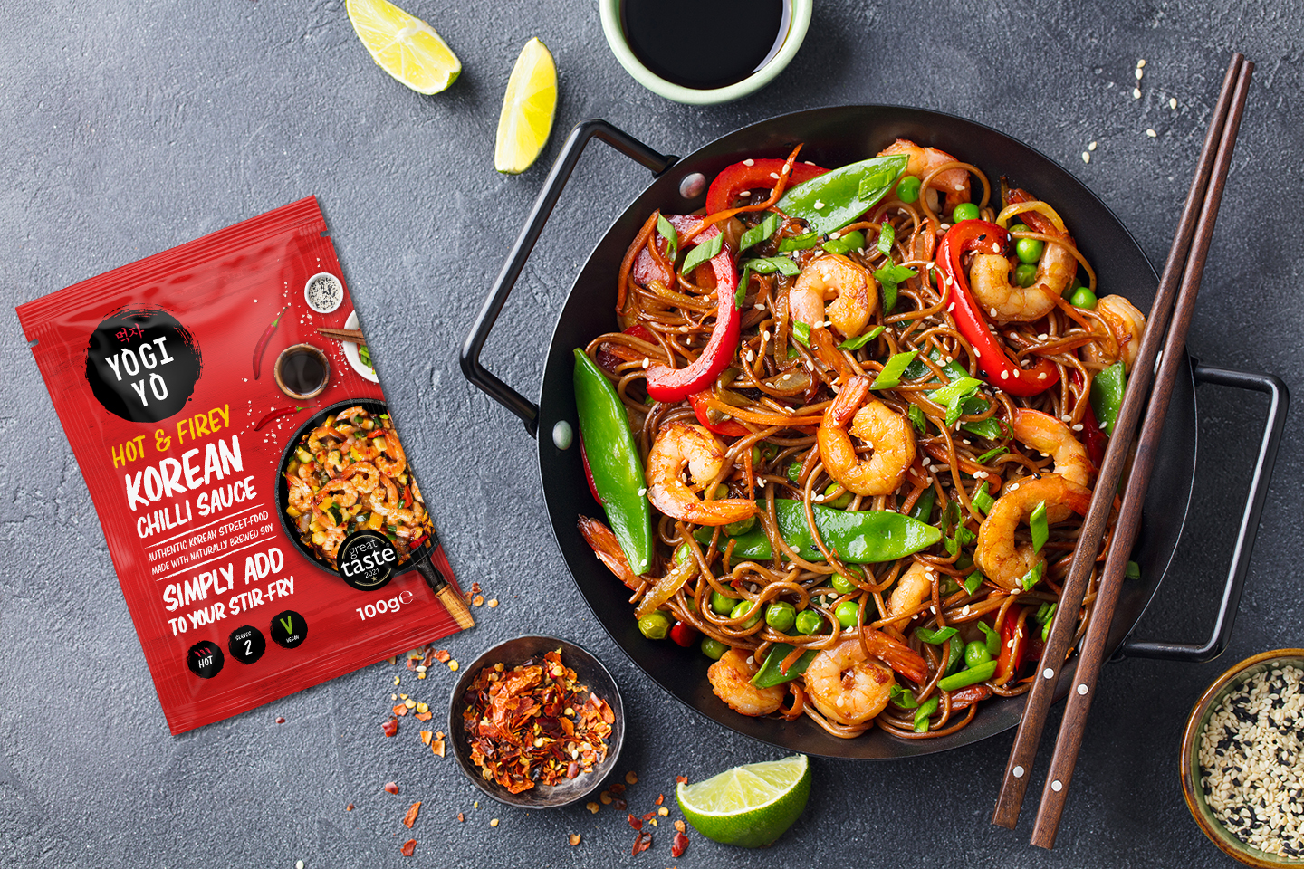 Black pan with shrimp stir-fry noodles, red bell peppers, snap peas, and green peas beside chopsticks and a packet of Yogi Yo hot and fiery Korean chili sauce.