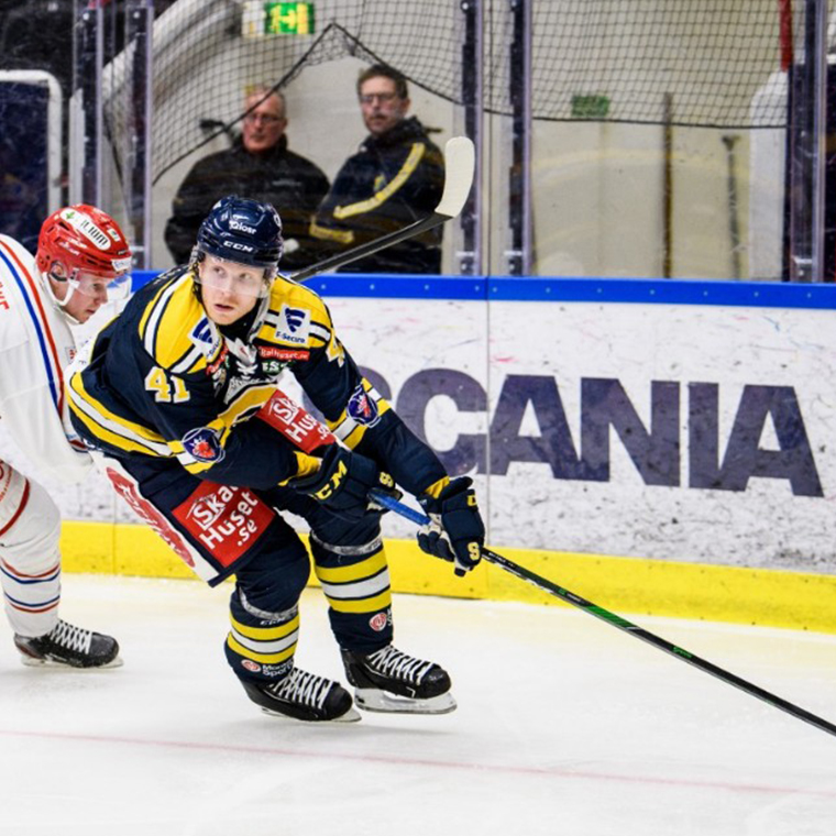 Two ice hockey players competing on the ice rink near the boards, one in yellow and navy and the other in white and red.
