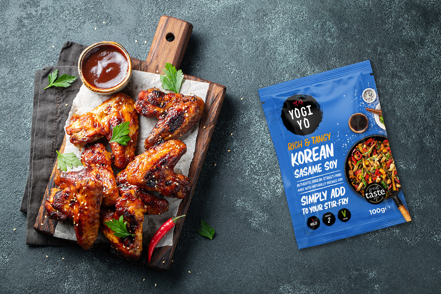Glazed chicken wings garnished with parsley on a wooden board with sauce, next to a packet of Yogi Yo Korean Sesame Soy sauce.