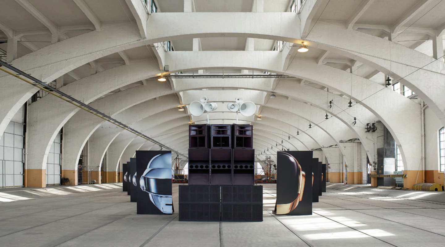 Large industrial warehouse with arched ceiling beams featuring a central sound system setup flanked by panels displaying helmets.