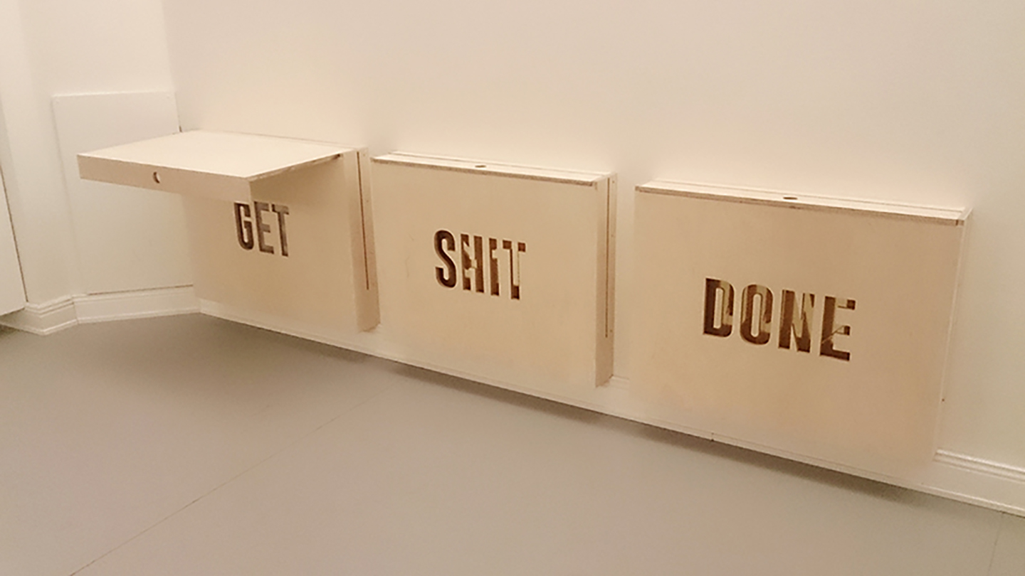 Three wall-mounted beige panels with the words GET, SHIT, and DONE cut out, with the first panel’s top flipped up like a shelf.