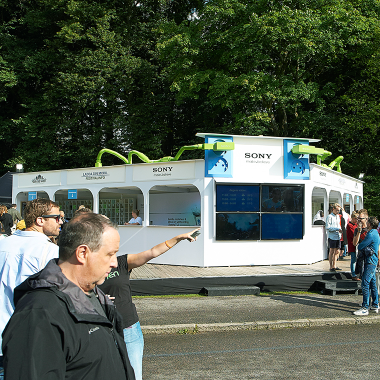 Outdoor Sony booth with digital screens and green decorations, surrounded by people at a festival in a park.