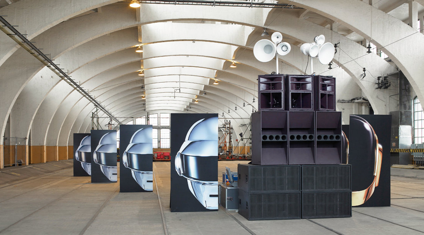 Large indoor space with arched ceiling showing a row of stands featuring Daft Punk helmets and stacked black speakers with white megaphones on top.