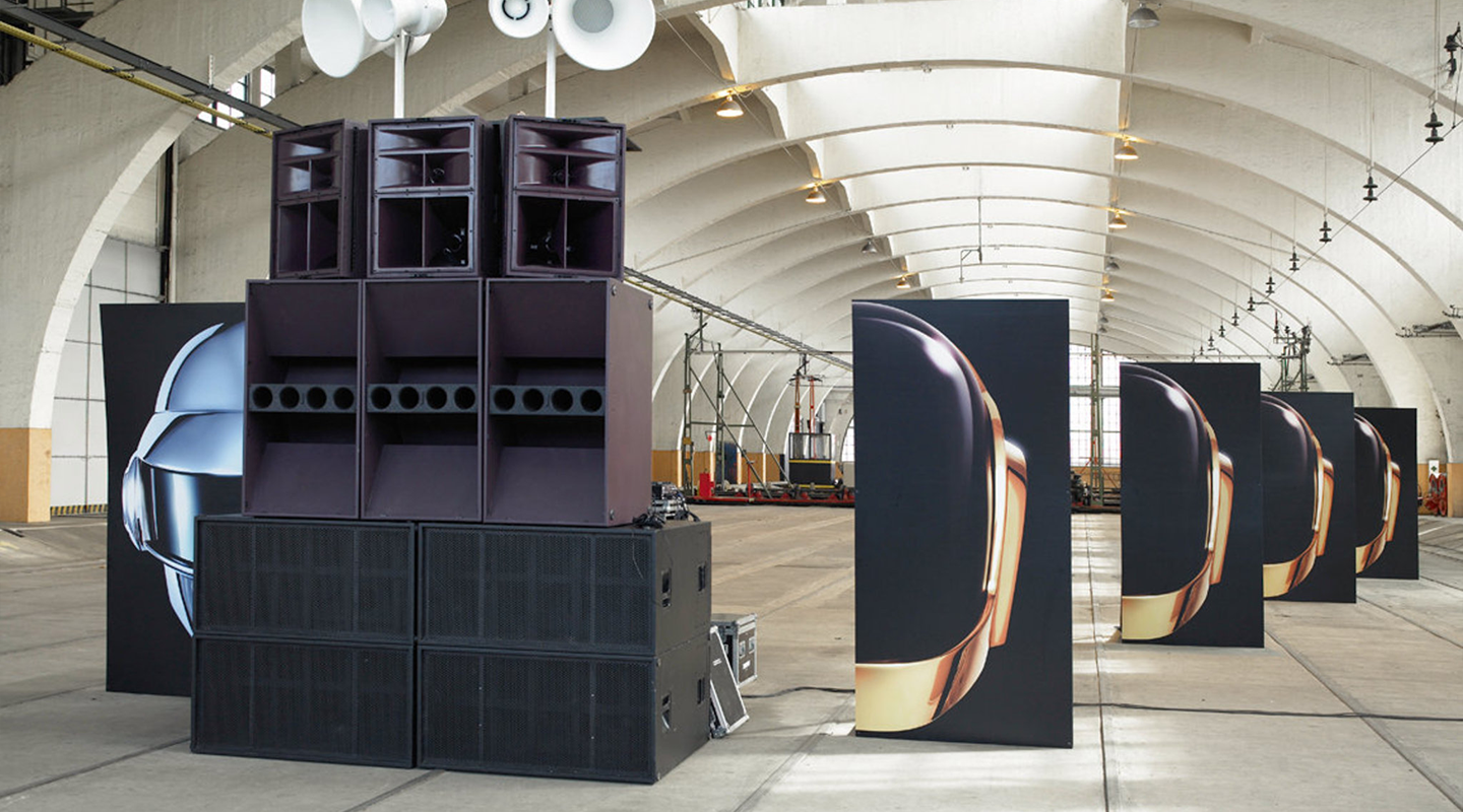 Large stacked speakers in a warehouse with large vertical panels displaying helmet images from the musical duo Daft Punk.