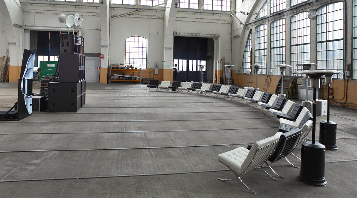 Curved row of black and white modern chairs in a large industrial-style room with tall windows and heaters.