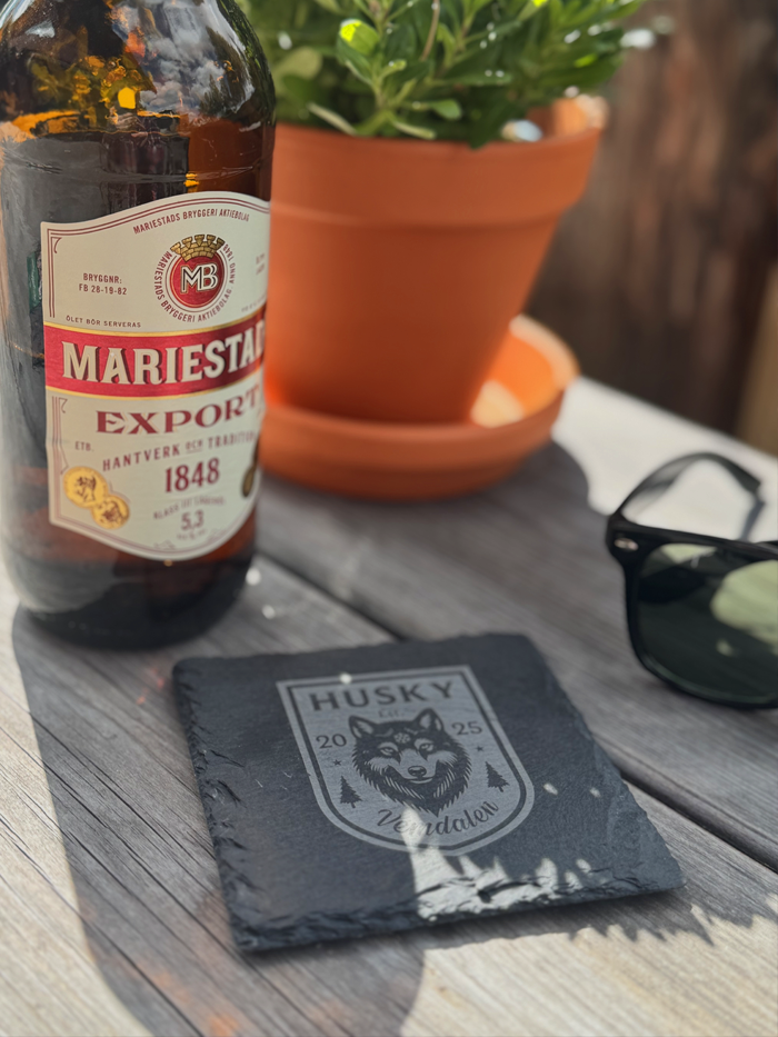 A Mariestads Export beer bottle next to a black coaster with a husky logo, a pair of sunglasses, and a potted plant on a wooden table.
