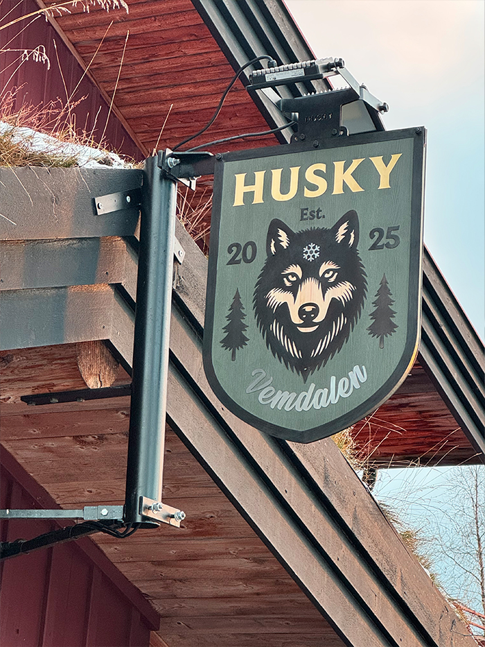 Sign with a husky dog face illustration and text reading 'HUSKY Est. 2025 Vemdalen' attached to a wooden building exterior.
