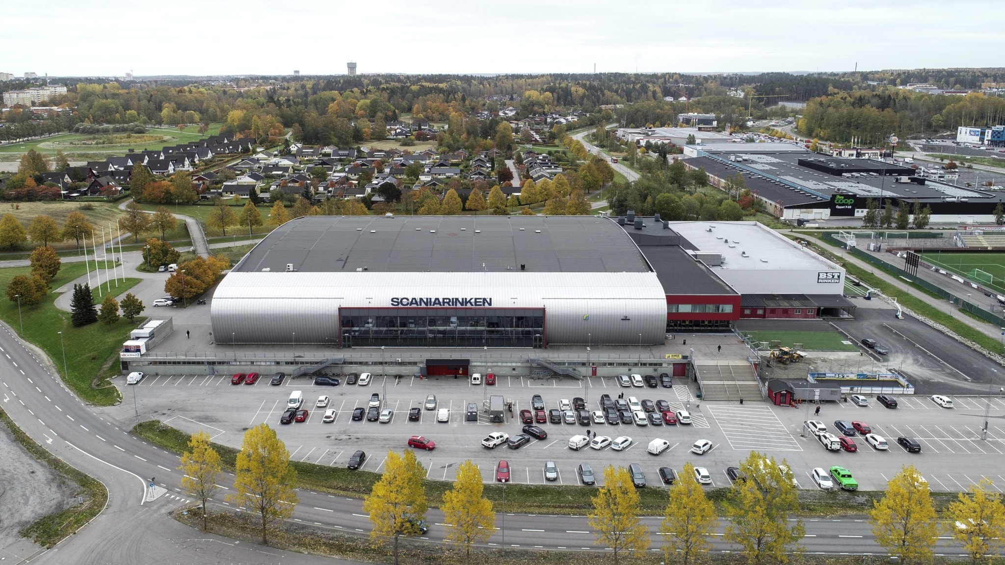 Aerial view of Scaniarinken sports arena with surrounding parking lot, yellow-leaved trees, and residential buildings in the background.