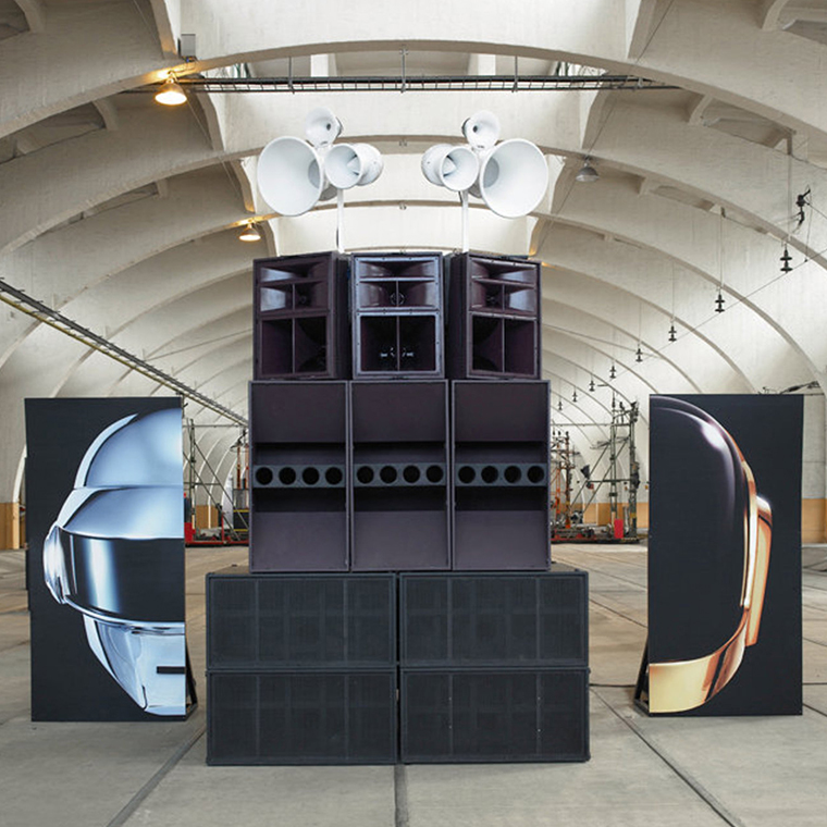 Large stacked black speakers with multiple white horn speakers on top, flanked by two vertical banners displaying stylized metallic helmets resembling Daft Punk's iconic headgear, in an industrial arched warehouse.