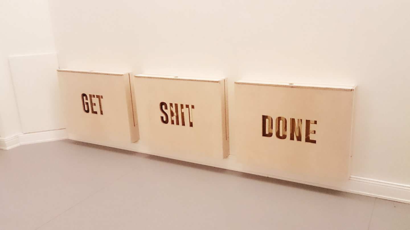 Three wooden panels leaning against a white wall with the words Get, Shit, Done cut out.