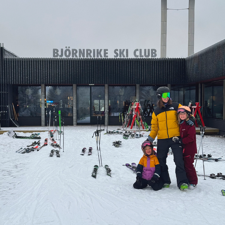 Three people wearing ski gear pose happily in front of the Björnrike Ski Club building, surrounded by skis and snow.