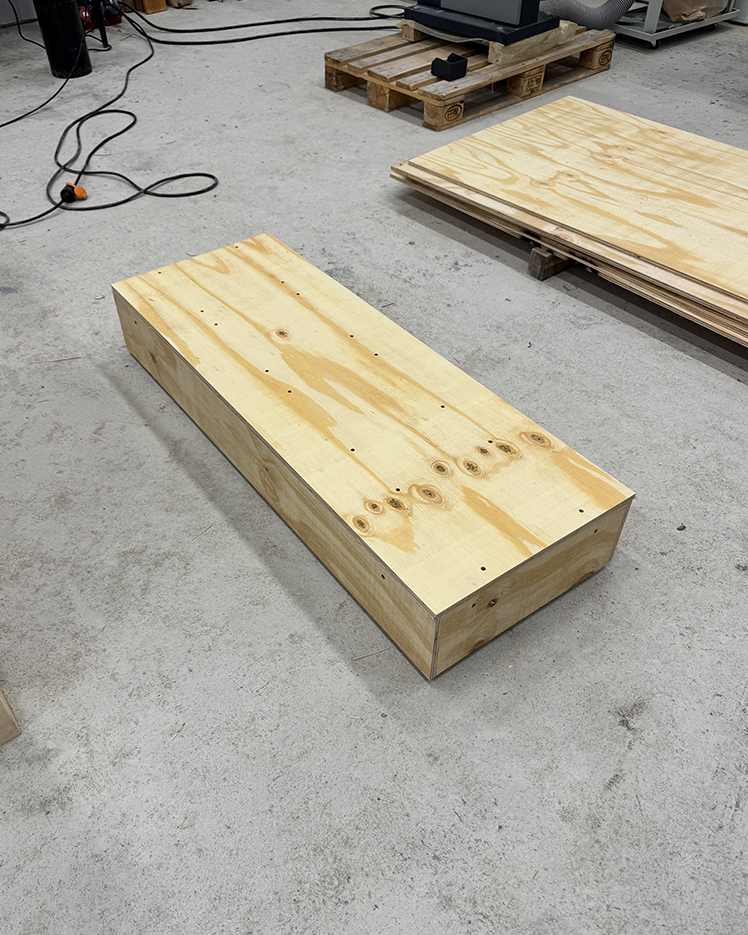 Rectangular wooden box with visible wood grain on a concrete floor, with wooden pallets and stacked plywood sheets in the background.