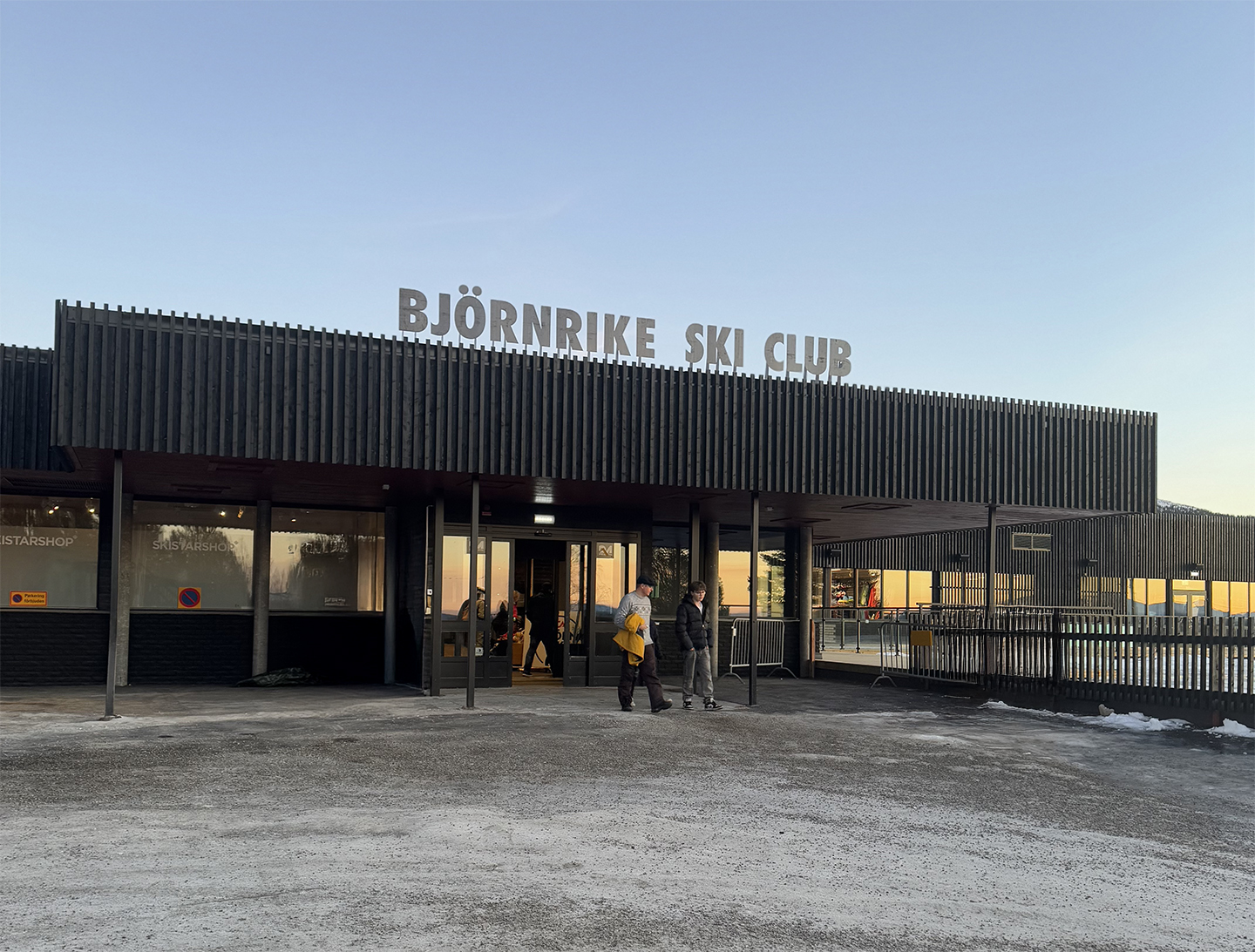 Exterior view of Björnrike Ski Club building with two people standing near the entrance during dusk.