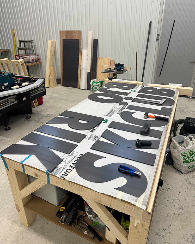 Workshop table with large black letters spelling 'SKI CLUB' laid out, surrounded by tools and wooden materials.