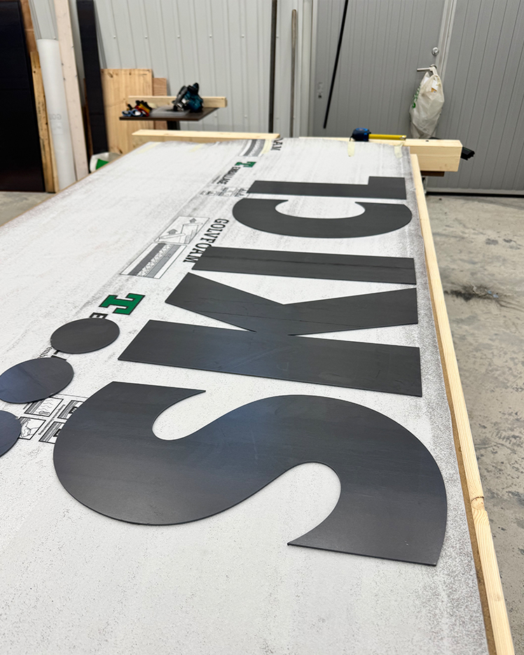 Large black letters spelling 'SKI CLUB' laid out on a white board in a workshop with tools and wooden frames in the background.