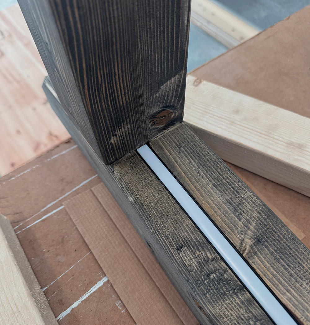 Close-up of a dark-stained wooden frame joint with a white LED strip embedded along the base wood piece, placed on a cardboard surface with lighter wooden boards around.
