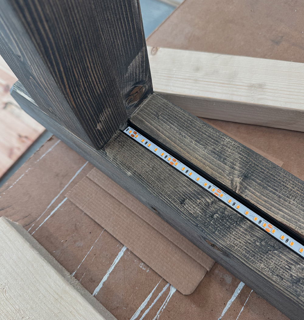 Close-up of a black wooden frame with an embedded LED strip light running along the inner edge, resting on a cardboard surface with additional wooden planks nearby.