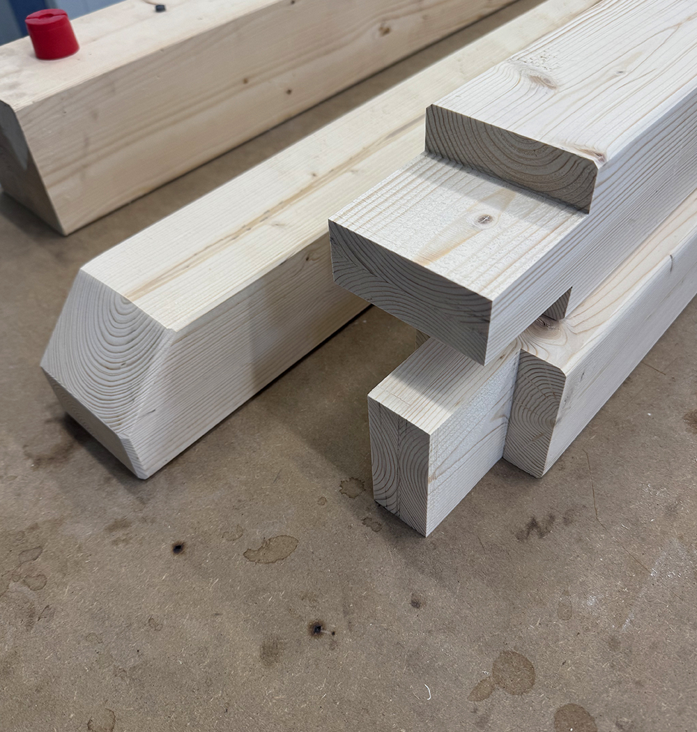 Stack of light-colored wooden beams, including one with a stepped mortise joint, placed on a workshop floor with a red cap in the background.