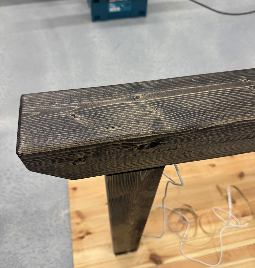 Close-up view of a dark-stained wooden beam with visible grain, knots, and textured surfaces, standing vertically on a lighter wood floor with a tangled white cable nearby.