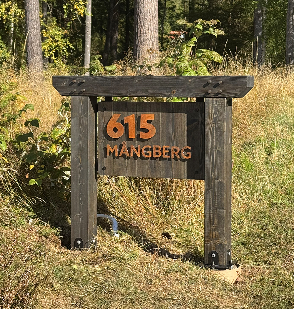 Rustic dark wooden sign with orange letters displaying house number '615' and the name 'MÅNGBERG' on grass with trees in the background.