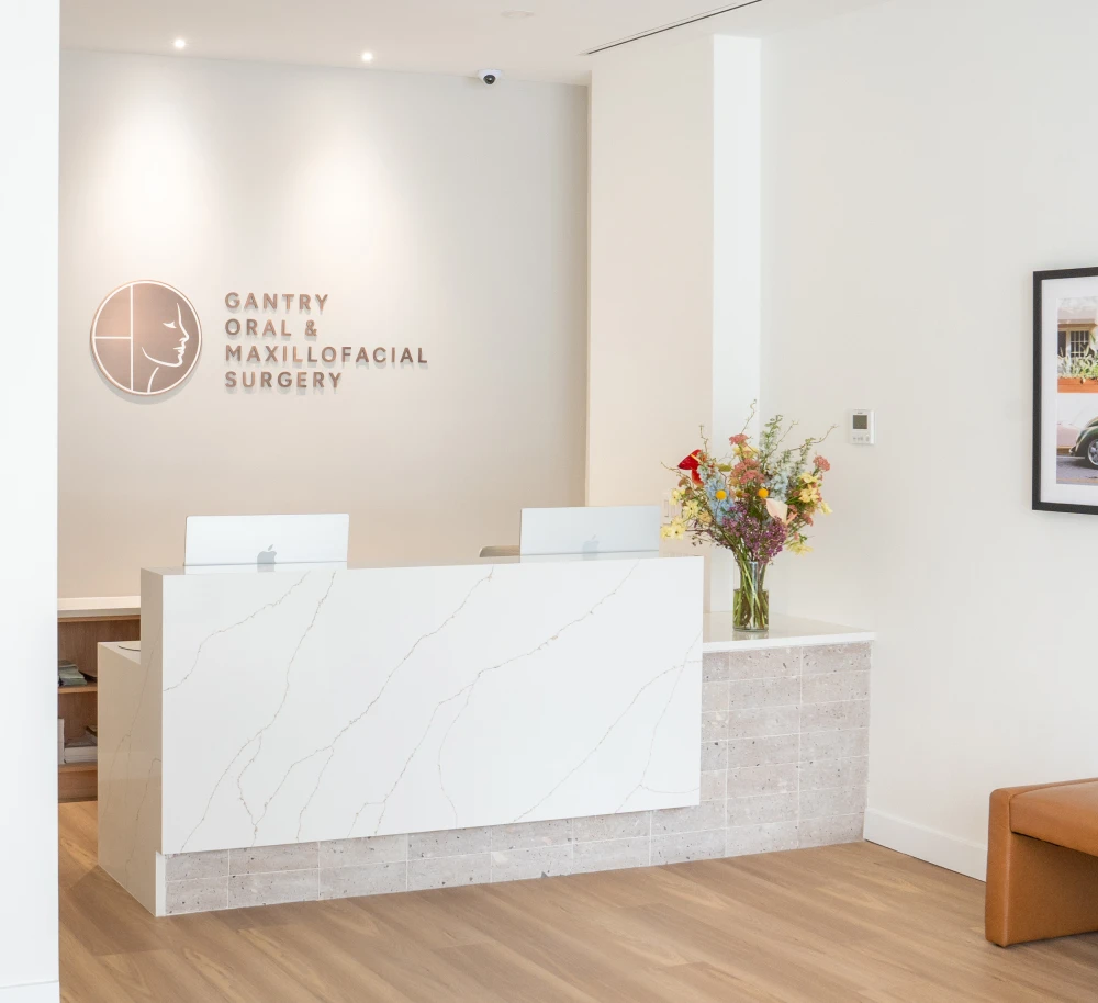 Reception area with white marble desk, two Apple computers, flower vase, and Gantry Oral & Maxillofacial Surgery sign.