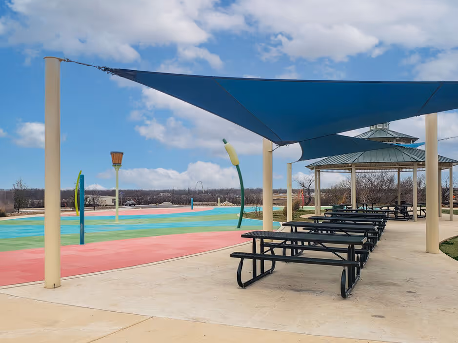Vibrant splash pad area with colorful ground and water features, alongside shaded picnic tables, at Enclave at Medina in Von Ormy, TX.