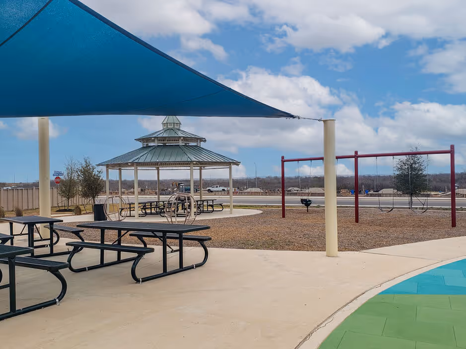Playground area with swings, a gazebo with picnic tables, and a large blue shade structure, providing family-friendly amenities at Enclave at Medina in Von Ormy, TX.