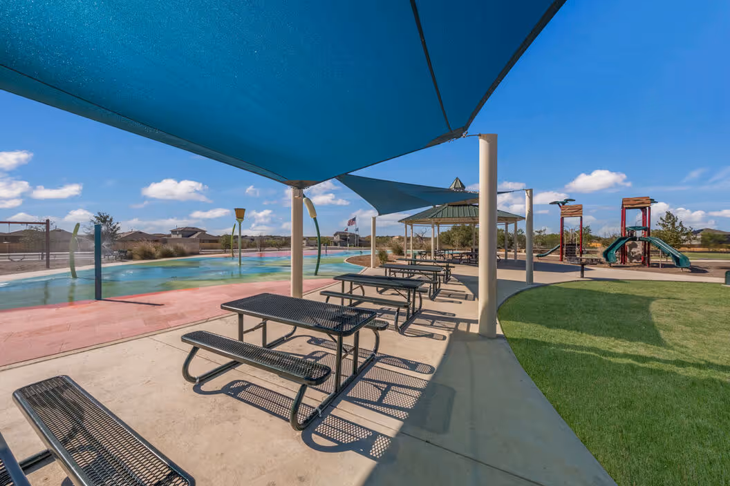 Covered outdoor picnic tables and a grassy area next to a colorful splash pad and playground, part of the community amenities at Enclave at Medina in Von Ormy, TX.