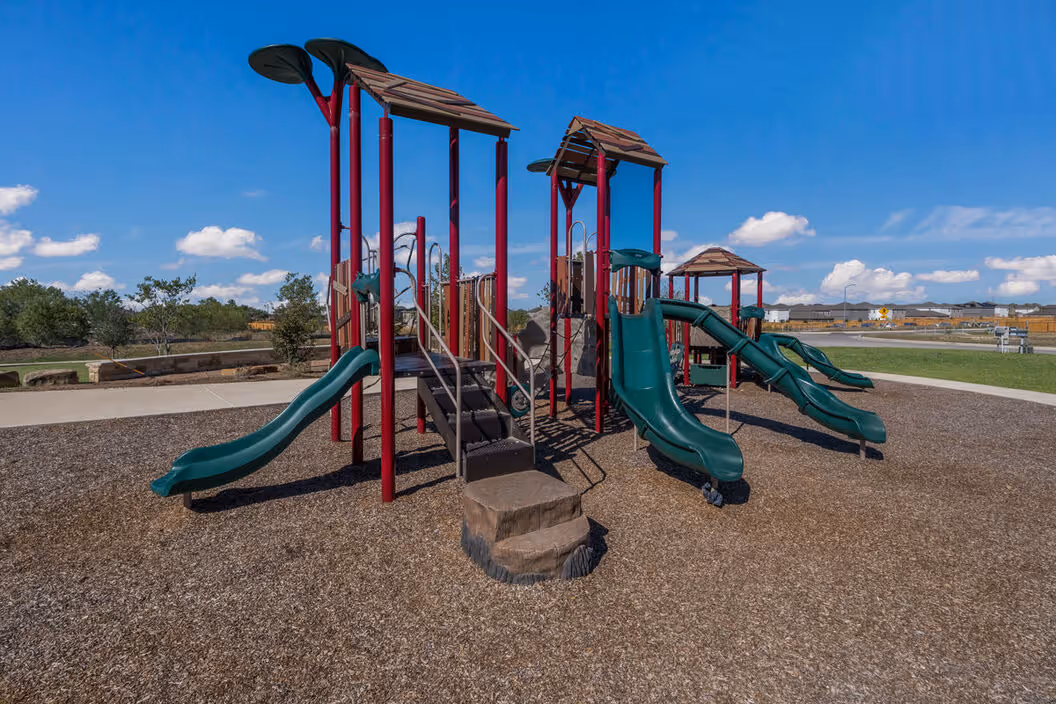 Large children's playground with multiple slides and climbing structures on wood chips, a fun amenity at Enclave at Medina in Von Ormy, TX.