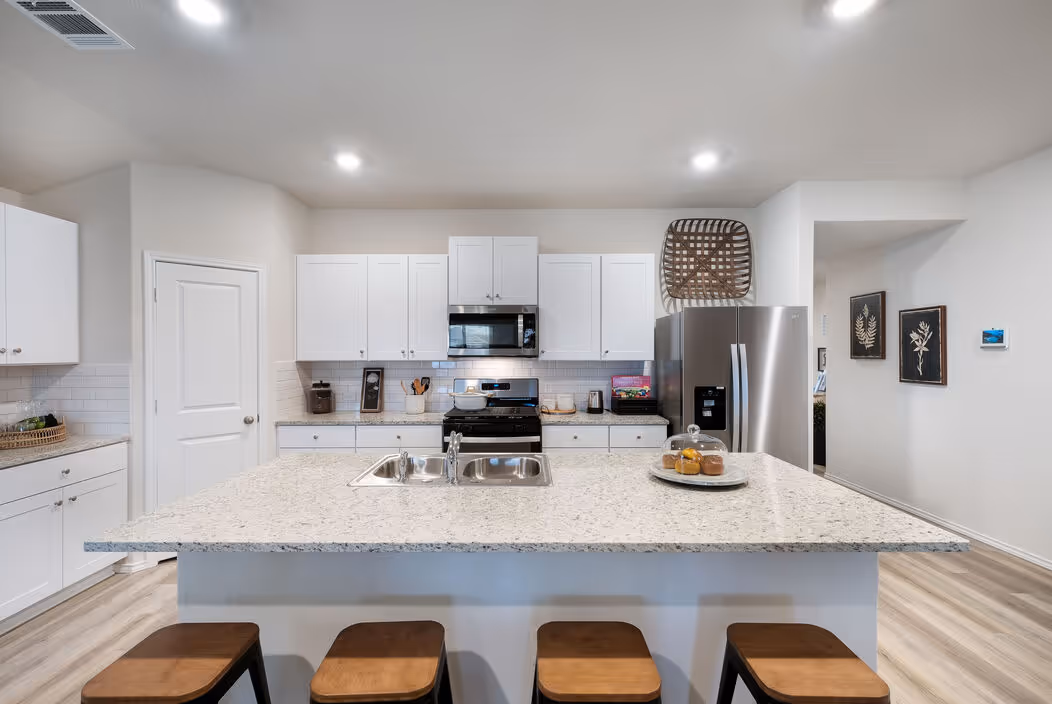 Bright and modern kitchen featuring white shaker cabinets, granite countertops, a large island with bar seating, and stainless steel appliances, in a home at Enclave at Medina in Von Ormy, TX.