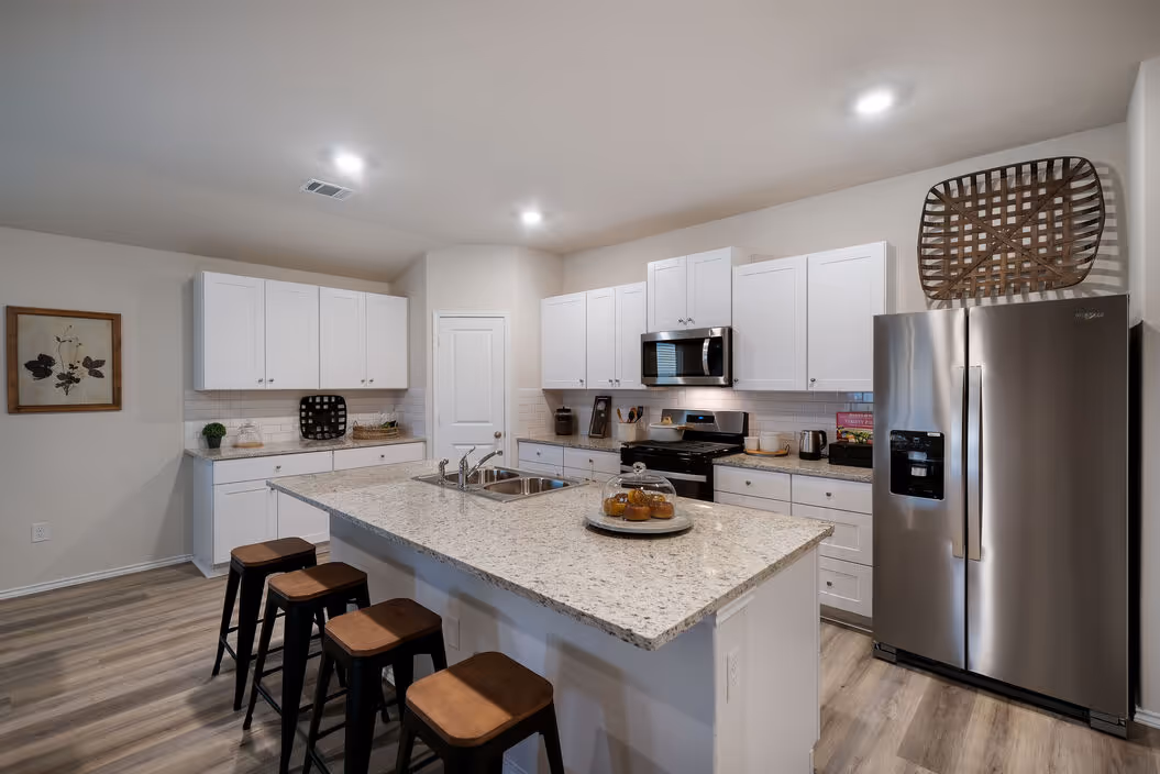 Bright kitchen featuring white shaker cabinets, granite countertops, and a large island with three bar stools, at Enclave at Medina in Von Ormy, TX.