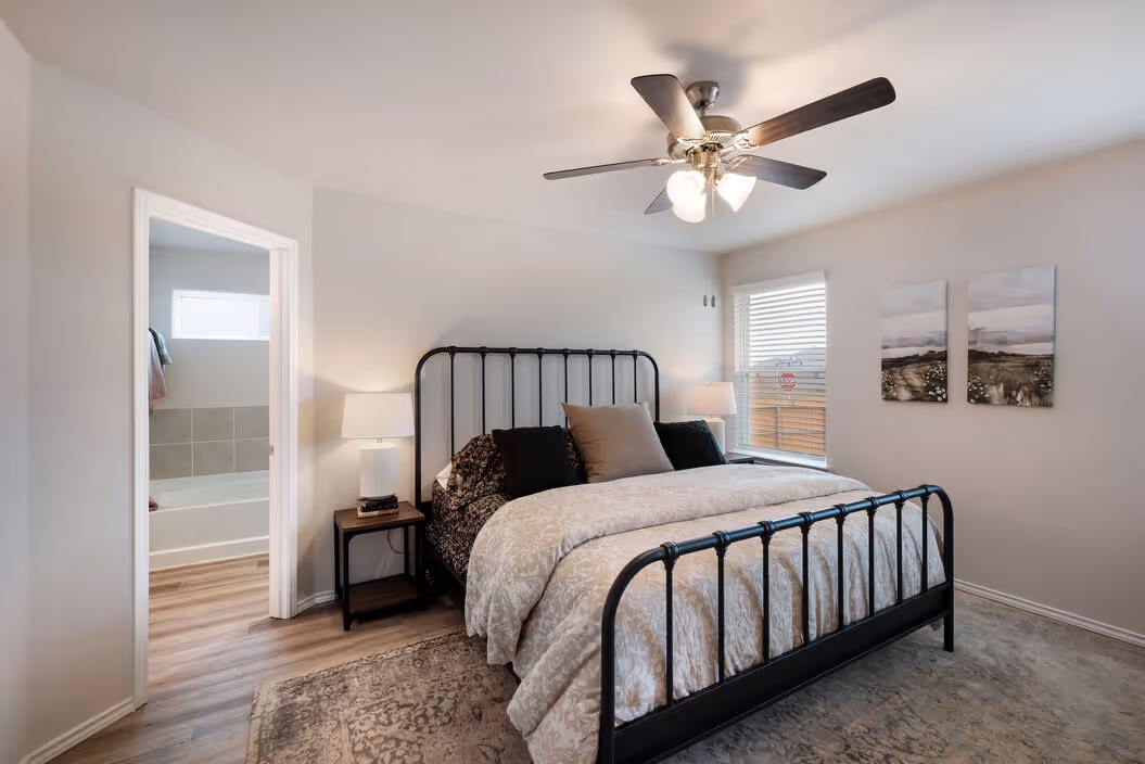 Master bedroom with a stylish black metal bed frame, ceiling fan, and en-suite bathroom access, in a home at Enclave at Medina in Von Ormy, TX.