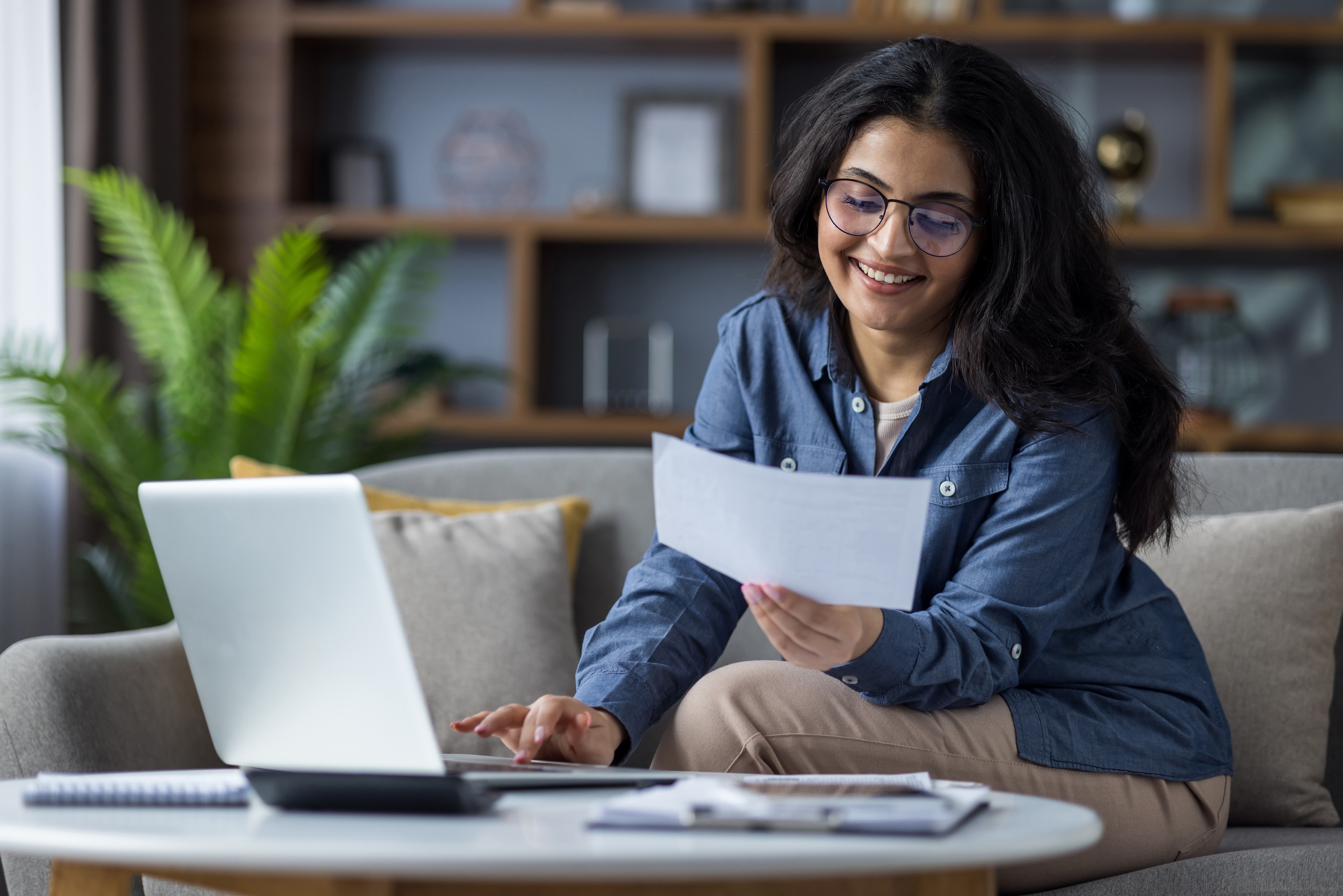 A smiling woman working from home on a laptop and reviewing documents on her sofa, representing a comfortable and convenient lifestyle at Enclave at Medina in Von Ormy, TX.