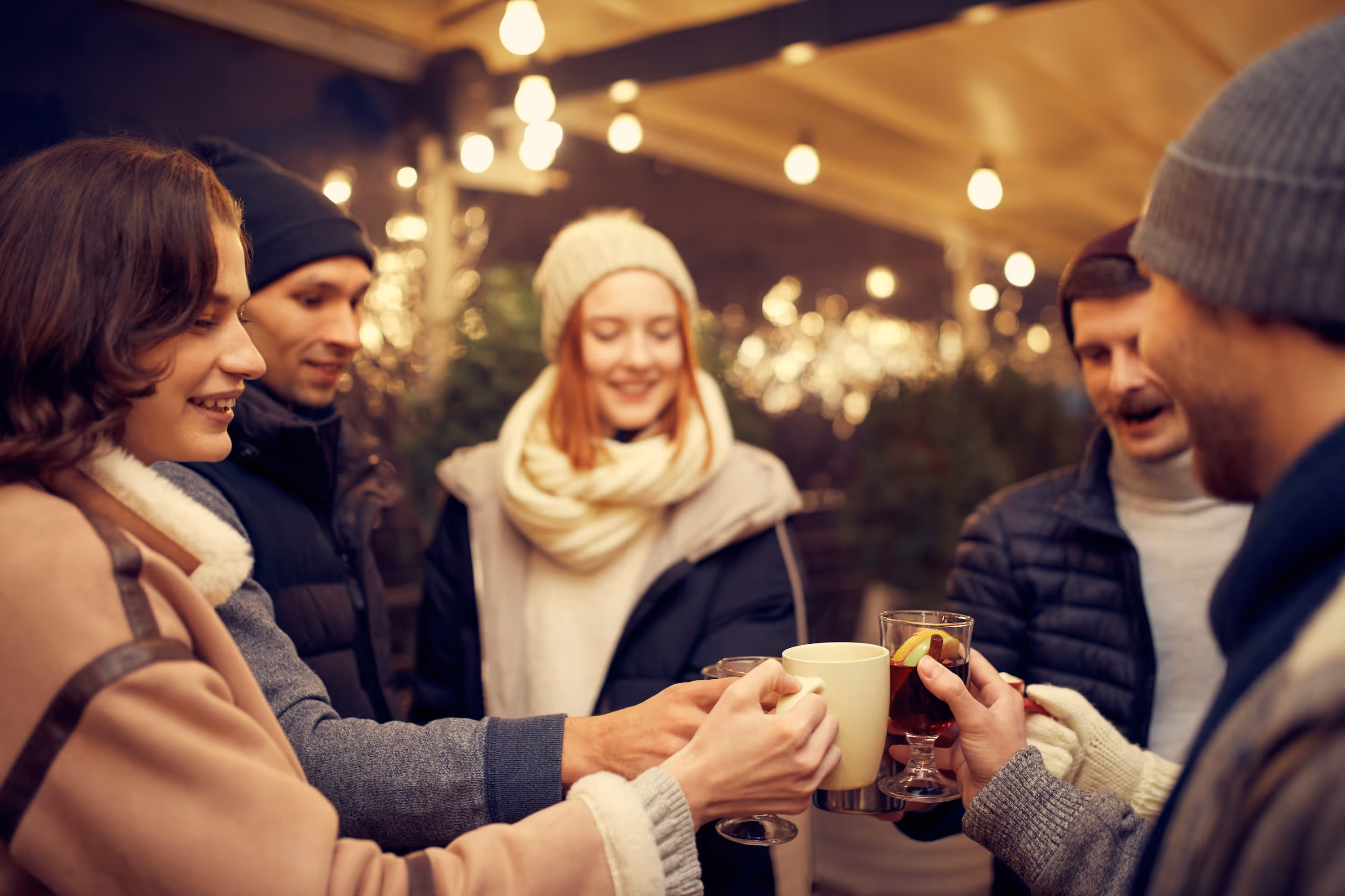 A group of smiling residents in winter hats and scarves clink mugs of warm beverages together under glowing outdoor string lights near Enclave at Medina.