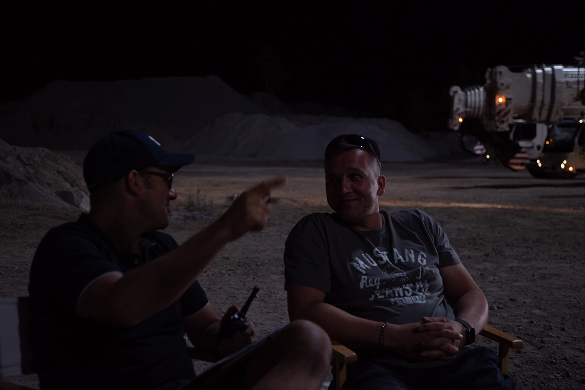 Imagefilm der Filmproduktion Panda Pictures aus München und Ulm für Maxikraft. Zwei Produktionsmitarbeiter unterhalten sich in entspannter Atmosphäre in der Nacht, mit Kieshaufen und einem LKW im Hintergrund, die für die Szene aufgebaut sind.